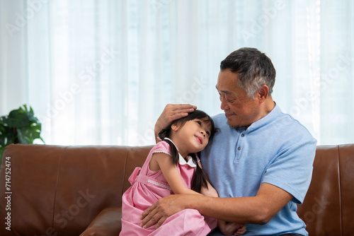 Happy Asian senior grandfather sits on a couch with his granddaughter and plays with his granddaughter in the living room at home, The Concept of family having fun in their house