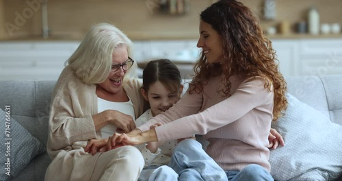 Cheerful grandma and mother tickling playful little kid girl on home sofa, playing with child, laughing, shouting, having fun, enjoying playtime, leisure together, hugging with love, care photo