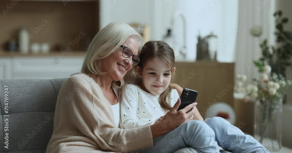 Cheerful loving granny and cute little granddaughter kid using smartphone on sofa together, typing on screen, using smart home application, virtual assistant, service, smiling, speaking, hugging Cheerful loving granny and cute little granddaughter kid using smartphone on sofa together, typing on screen, using smart home application, virtual assistant, service, smiling, speaking, hugging