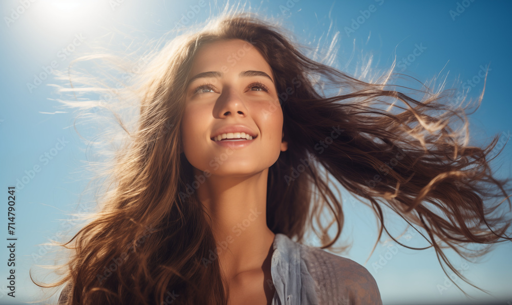 portret młodej brunetki z długimi włosami cieszącej się jasnym słońcem na tle błękitnego nieba - obrazy, fototapety, plakaty 