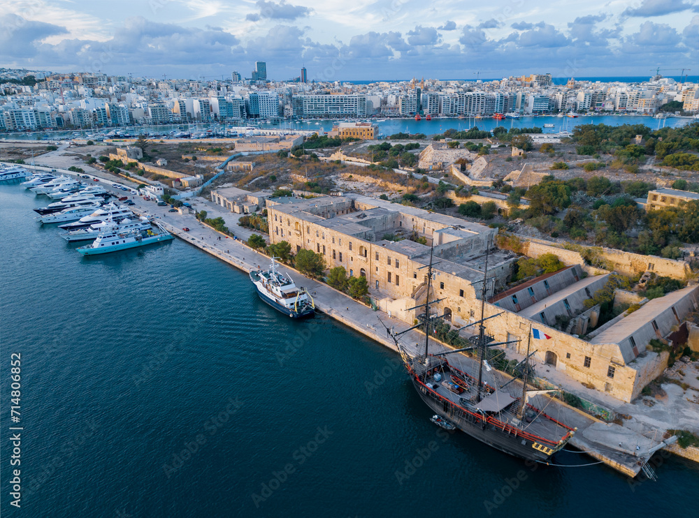 Naklejka premium Beautiful sunset on Lazaretto of Manoel Island with Yacht Marina and old wooden sailing ship with French flag. Aerial view on a city skyline with natural golden and blue colors. Clouds in the sky.