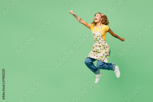 Full body side view young housewife housekeeper chef cook baker woman wear apron yellow t-shirt jump high do super hero gesture pov fly isolated on plain green background studio. Cooking food concept.