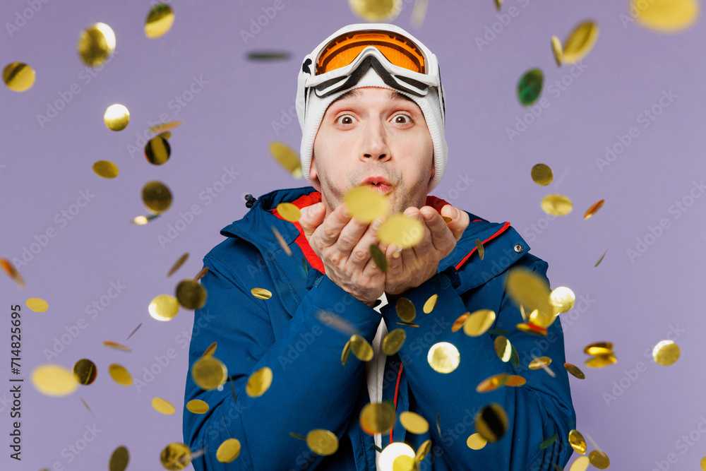 Skier Caucasian man he wears warm blue windbreaker jacket ski goggles mask hat blow confetti glitters look camera spend extreme weekend winter season in mountains isolated on plain purple background. Skier Caucasian man he wears warm blue windbreaker jacket ski goggles mask hat blow confetti glitters look camera spend extreme weekend winter season in mountains isolated on plain purple background.