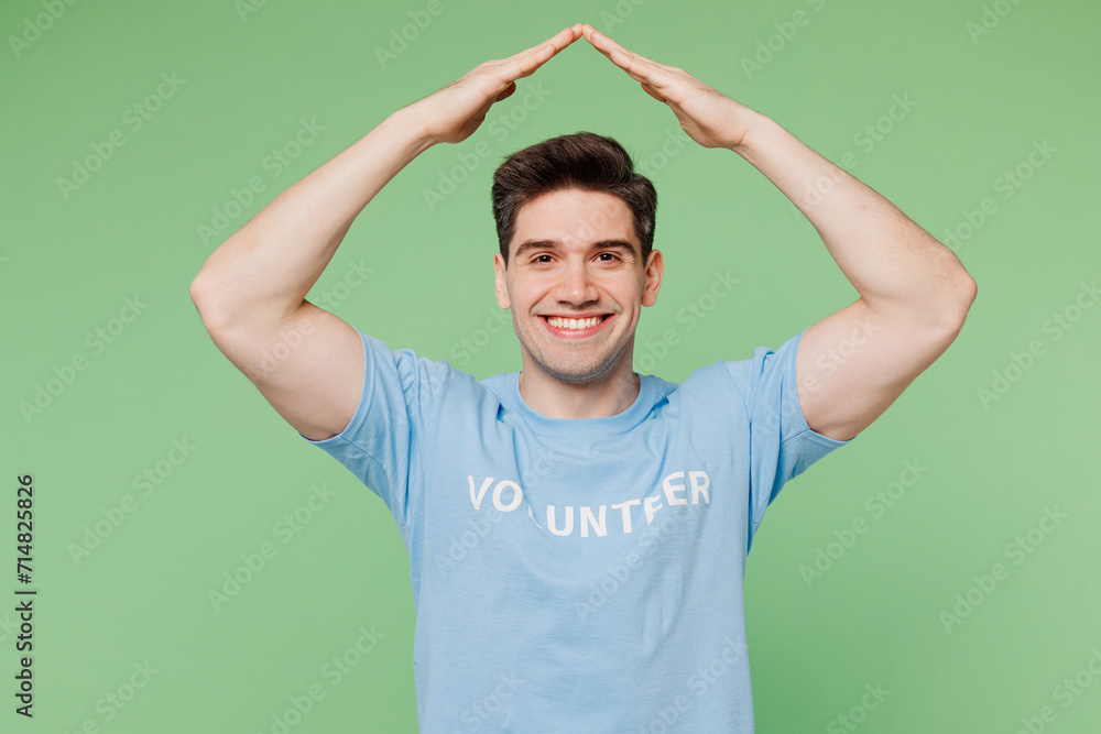 Fototapeta premium Young man he wear blue t-shirt white title volunteer hold folded hands above head like house roof isolated on plain pastel green background. Voluntary free work assistance help charity grace concept.