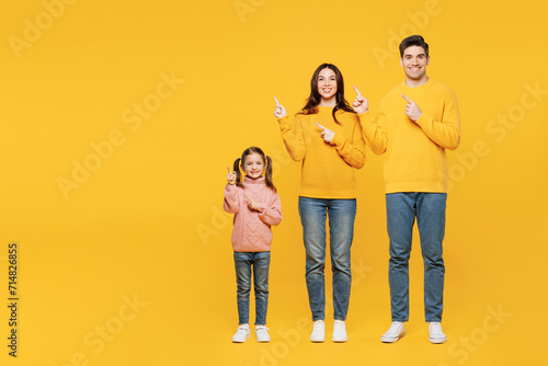Full body young fun happy parents mom dad with child kid girl 7-8 years old wear pink sweater casual clothes point index finger aside on area isolated on plain yellow background. Family day concept.
