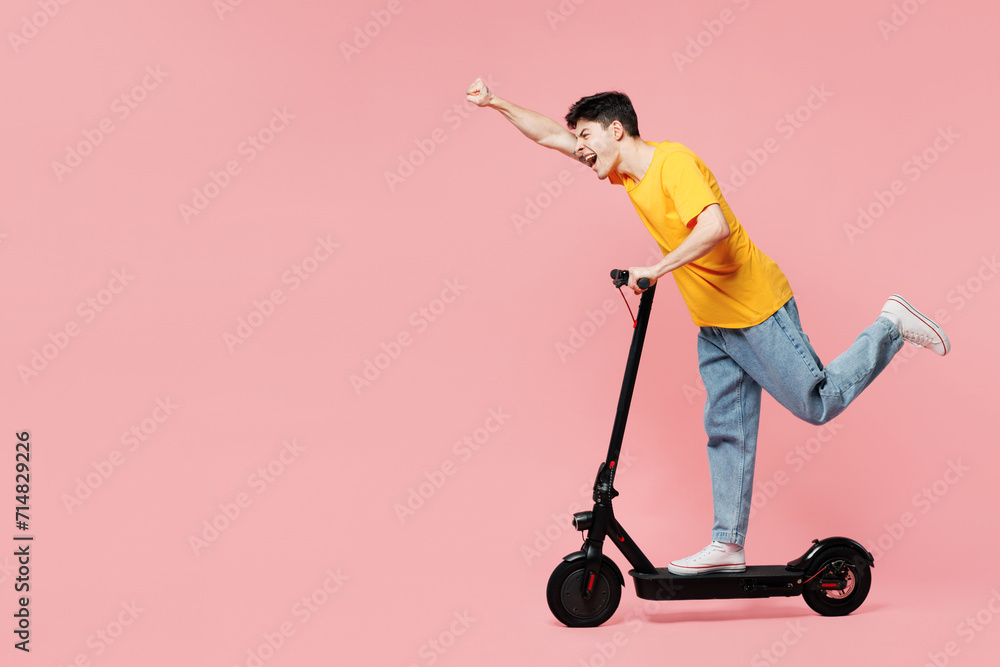 Full body side profile view student happy young man he wears yellow t-shirt casual clothes driving electric scooter isolated on plain pastel light pink background studio portrait. Lifestyle concept. Full body side profile view student happy young man he wears yellow t-shirt casual clothes driving electric scooter isolated on plain pastel light pink background studio portrait. Lifestyle concept.