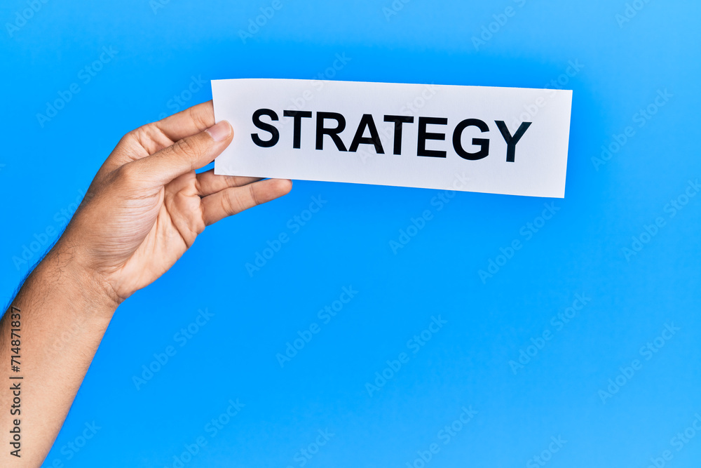 Hand of caucasian man holding paper with strategy word over isolated blue background Hand of caucasian man holding paper with strategy word over isolated blue background