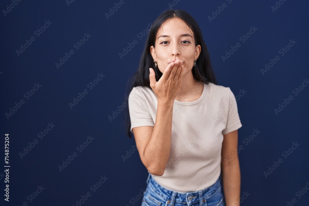 Young hispanic woman standing over blue background looking at the camera blowing a kiss with hand on air being lovely and sexy. love expression. Young hispanic woman standing over blue background looking at the camera blowing a kiss with hand on air being lovely and sexy. love expression.