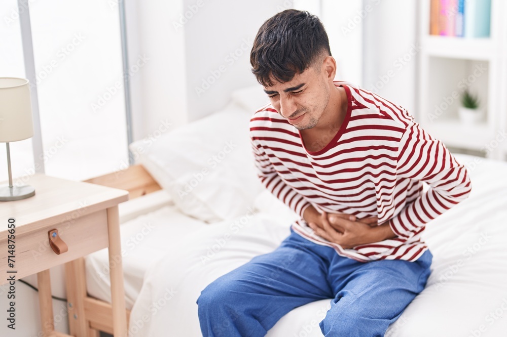 Young hispanic man suffering for stomach ache sitting on bed at bedroom