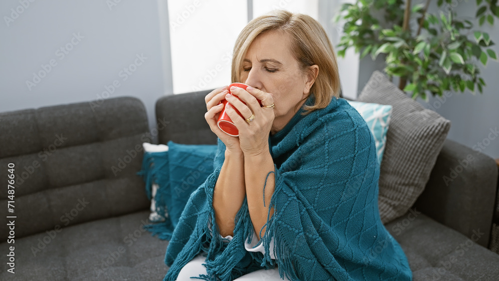 A mature woman enjoys a hot beverage while wrapped in a cozy throw, sitting on her sofa at home. A mature woman enjoys a hot beverage while wrapped in a cozy throw, sitting on her sofa at home.