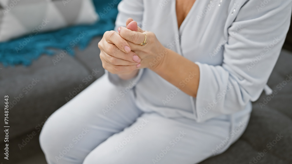 A middle-aged caucasian woman dressed in white sits indoors, clasping hands expressing contemplation or calmness. A middle-aged caucasian woman dressed in white sits indoors, clasping hands expressing contemplation or calmness.