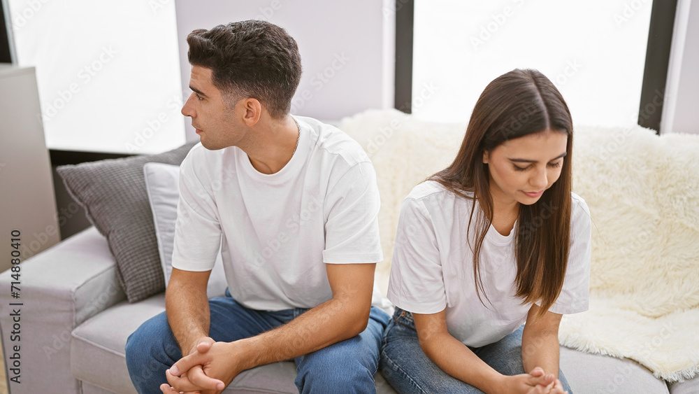 A man and a woman sit apart on a couch, suggesting tension or disagreement in a modern living room setting. A man and a woman sit apart on a couch, suggesting tension or disagreement in a modern living room setting.