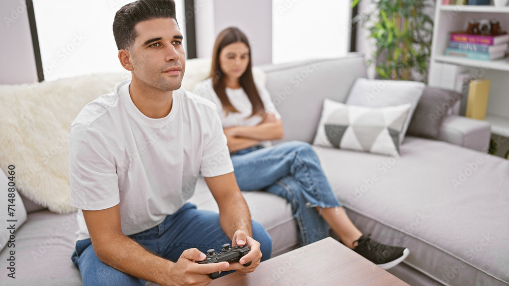 A man plays video games while a woman sits behind him, both on a couch in a modern living room setting. A man plays video games while a woman sits behind him, both on a couch in a modern living room setting.