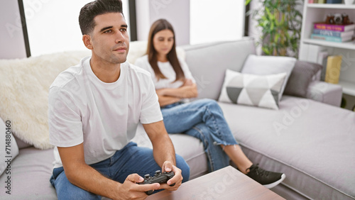 A man plays video games while a woman sits behind him, both on a couch in a modern living room setting.
