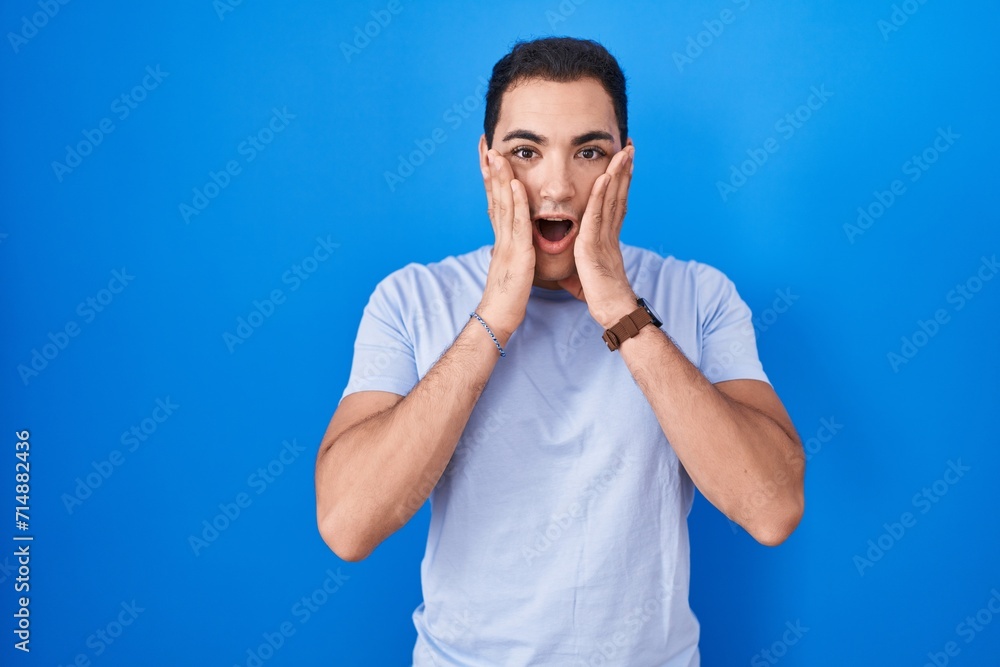 Young hispanic man standing over blue background afraid and shocked, surprise and amazed expression with hands on face Young hispanic man standing over blue background afraid and shocked, surprise and amazed expression with hands on face