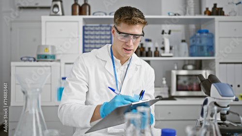 Handsome young caucasian scientist man  smiling as he writes on clipboard at bustling medical lab