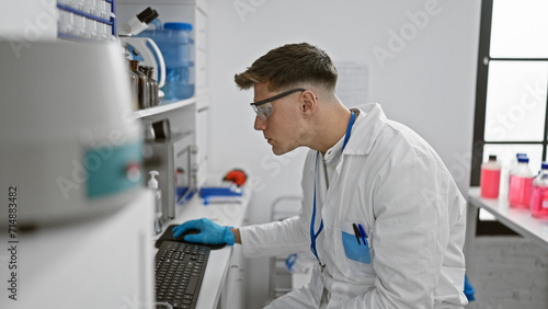 Attractive young caucasian male scientist totally engrossed in typing on computer amidst test tubes and microscopes, indoor laboratory work