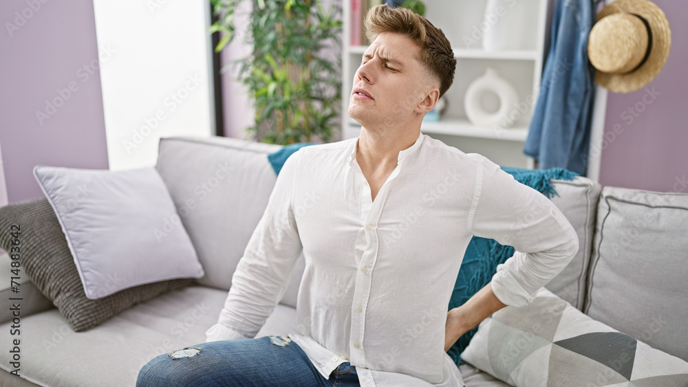 Worried young caucasian man grimacing in serious back pain, suffering, sitting on the living room sofa, in the background of his home, injured and unhappy from the backache Worried young caucasian man grimacing in serious back pain, suffering, sitting on the living room sofa, in the background of his home, injured and unhappy from the backache