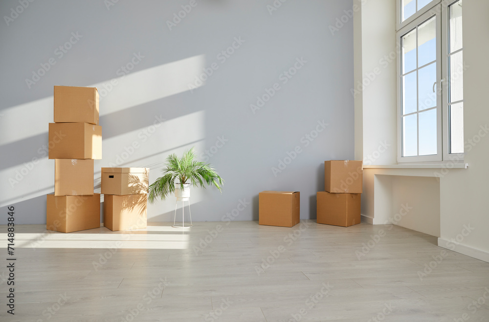 Unpacked cardboard boxes with things and a green plant are stacked on the floor in the empty living room in new apartment. Relocating, moving, delivery, real estate or mortgage concept. Unpacked cardboard boxes with things and a green plant are stacked on the floor in the empty living room in new apartment. Relocating, moving, delivery, real estate or mortgage concept.