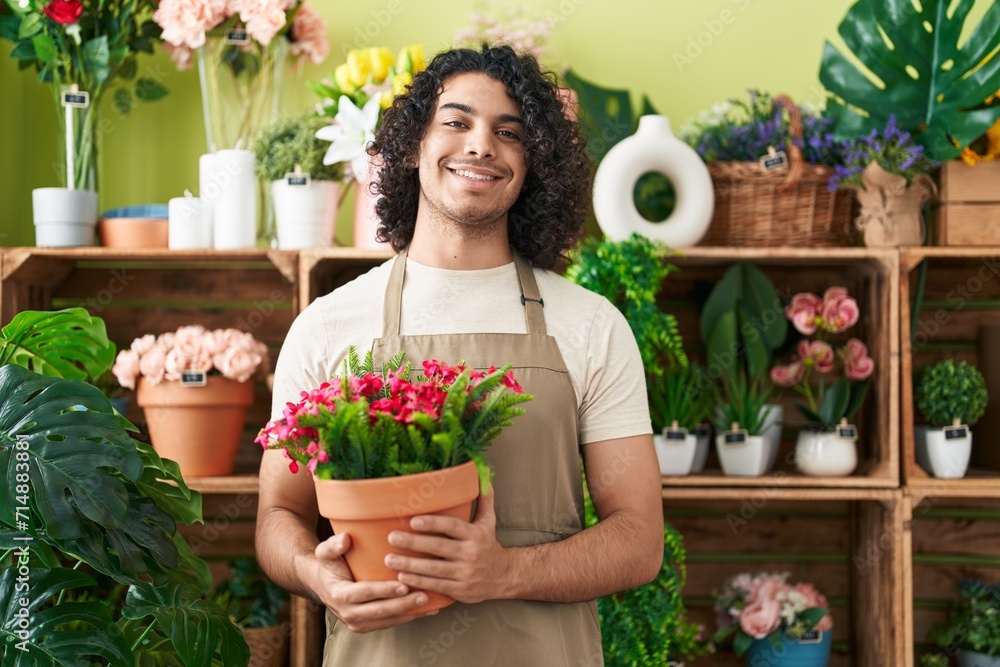 Hispanic man with curly hair working at florist shop holding plant looking positive and happy standing and smiling with a confident smile showing teeth