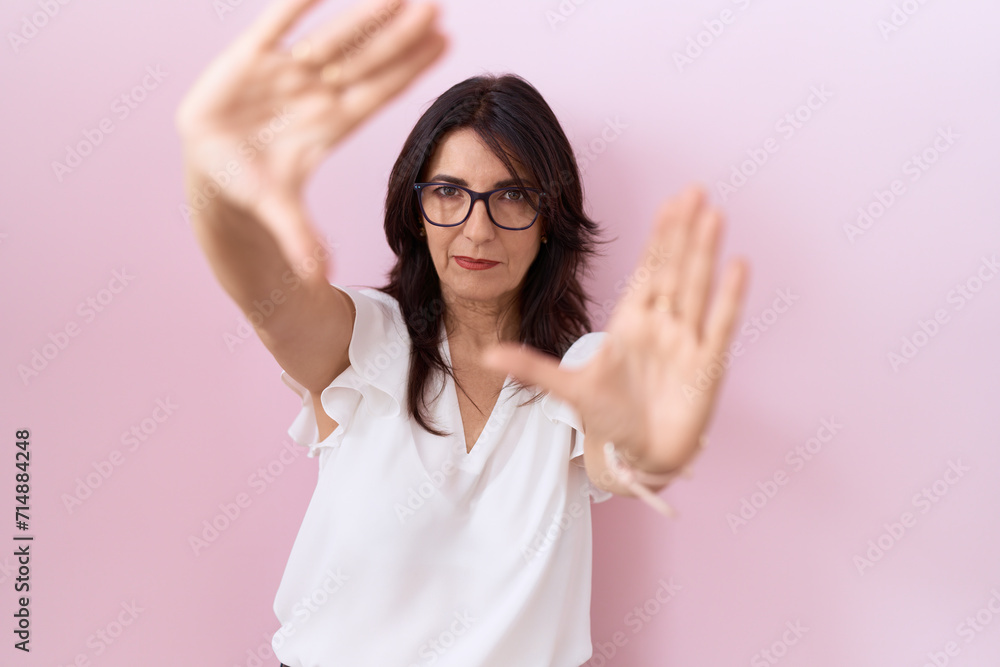 Fototapeta premium Middle age hispanic woman wearing casual white t shirt and glasses doing frame using hands palms and fingers, camera perspective