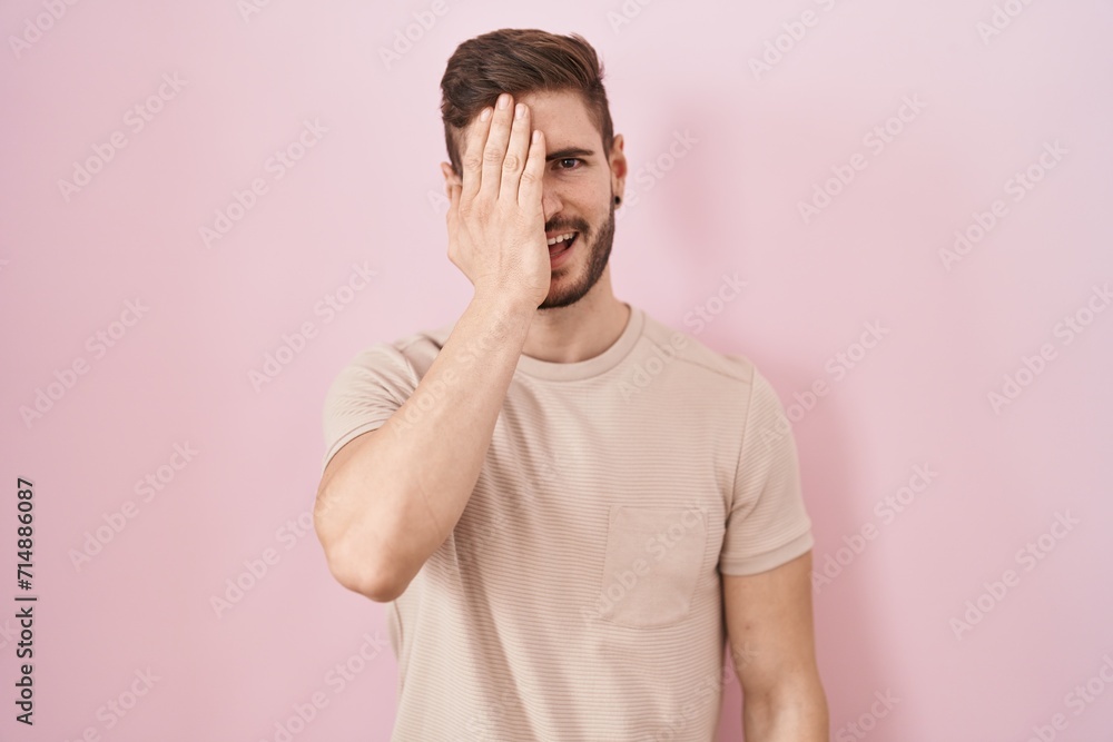 Hispanic man with beard standing over pink background covering one eye with hand, confident smile on face and surprise emotion. Hispanic man with beard standing over pink background covering one eye with hand, confident smile on face and surprise emotion.