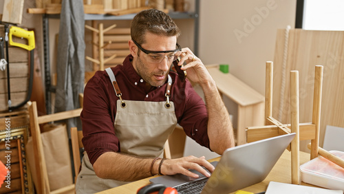 Handsome hispanic man with beard wearing glasses and apron uses laptop and phone in carpentry workshop.