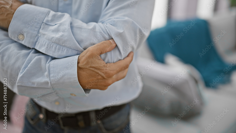 Cool, handsome middle age hispanic man suffering elbow joint pain standing in his indoor apartment living room - mature adult male experiencing bone ache problem at home Cool, handsome middle age hispanic man suffering elbow joint pain standing in his indoor apartment living room - mature adult male experiencing bone ache problem at home