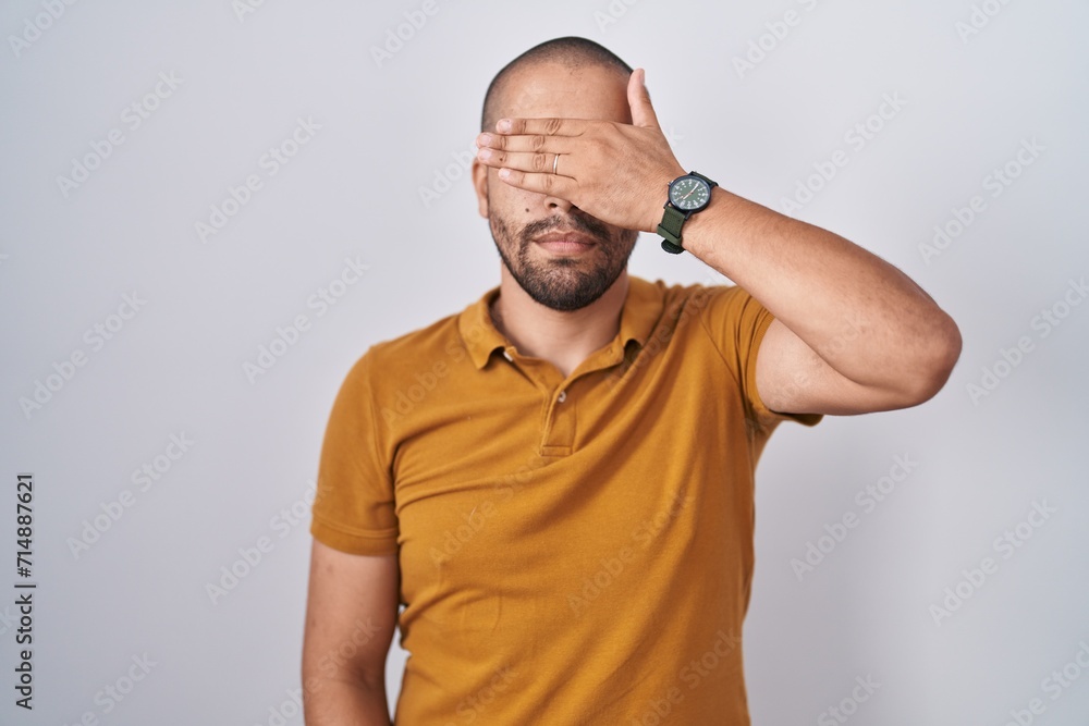 Hispanic man with beard standing over white background covering eyes with hand, looking serious and sad. sightless, hiding and rejection concept Hispanic man with beard standing over white background covering eyes with hand, looking serious and sad. sightless, hiding and rejection concept