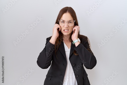 Beautiful brunette woman wearing business jacket and glasses covering ears with fingers with annoyed expression for the noise of loud music. deaf concept.