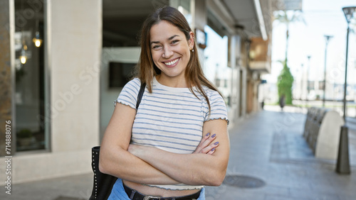 A smiling young woman with long brunette hair and blue eyes stands outdoors in a city street, arms crossed. photo