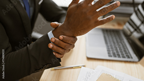 African man experiences wrist pain while working in the office environment, with a laptop and documents in the background. photo