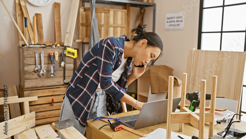 Focused asian woman working on a laptop in a well-equipped carpentry workshop