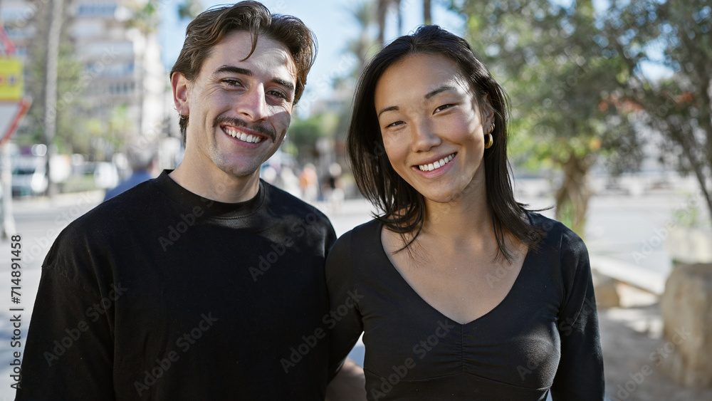 A smiling interracial couple, a man and a woman, standing together on a sunny urban street.