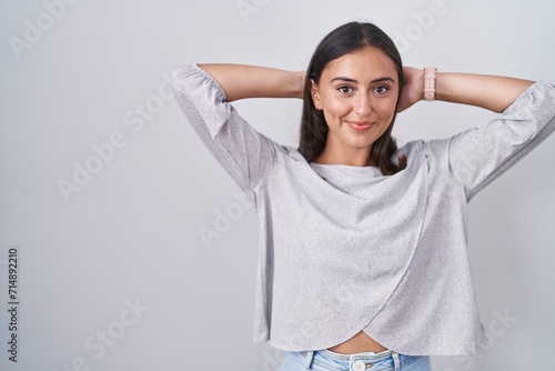 Young hispanic woman standing over white background relaxing and stretching, arms and hands behind head and neck smiling happy © Krakenimages.com