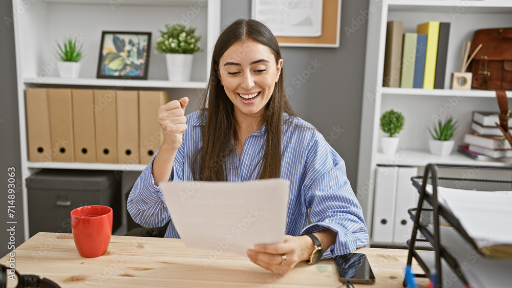 Hispanic woman excitedly reading document in modern office setting. Hispanic woman excitedly reading document in modern office setting.