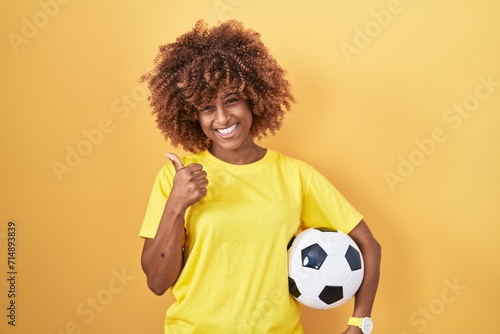 Young hispanic woman with curly hair holding football ball smiling happy and positive, thumb up doing excellent and approval sign