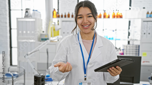 A smiling young woman in a lab coat holding a tablet stands in a pharmacy with shelves of medication in the background.