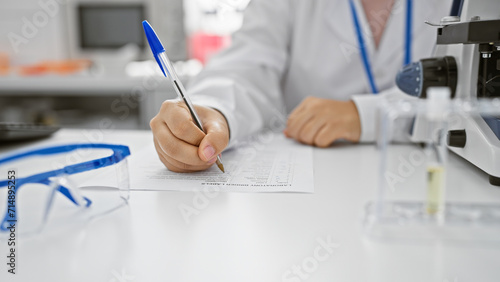 Professional woman in lab coat writing on a form, with microscope and eyeglasses in a clinical laboratory setting.