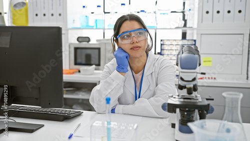 A contemplative hispanic woman scientist in lab coat and safety glasses sits in a laboratory with microscope and computer.