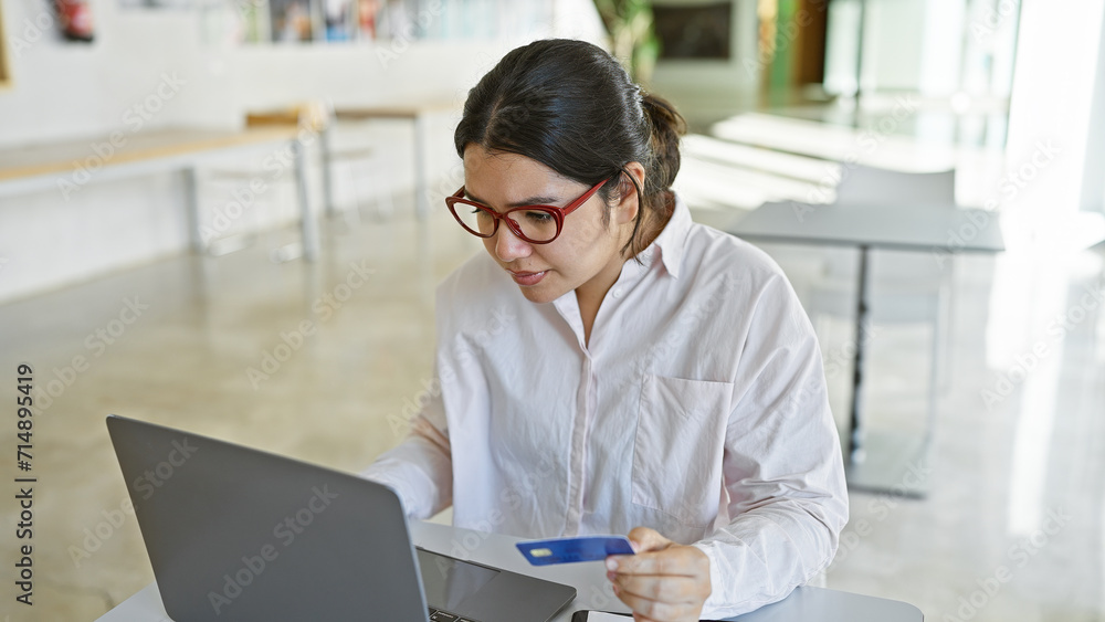 A focused hispanic woman in an office holding a credit card while working on a laptop, embodying professionalism and finance. A focused hispanic woman in an office holding a credit card while working on a laptop, embodying professionalism and finance.