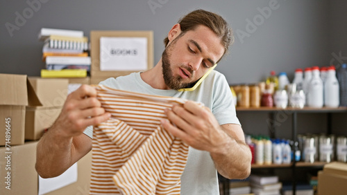 Young hispanic man volunteer packing clothes on cardboard box talking on smartphone at charity center