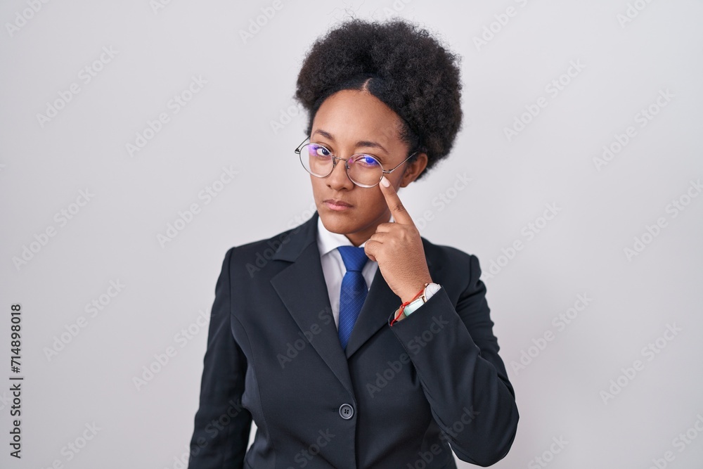 Beautiful african woman with curly hair wearing business jacket and glasses pointing to the eye watching you gesture, suspicious expression