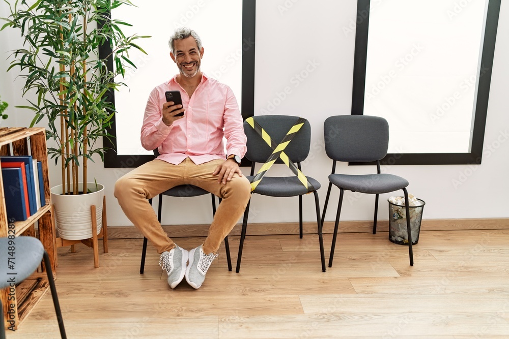Middle age grey-haired man using smartphone sitting on chair at waiting room Middle age grey-haired man using smartphone sitting on chair at waiting room