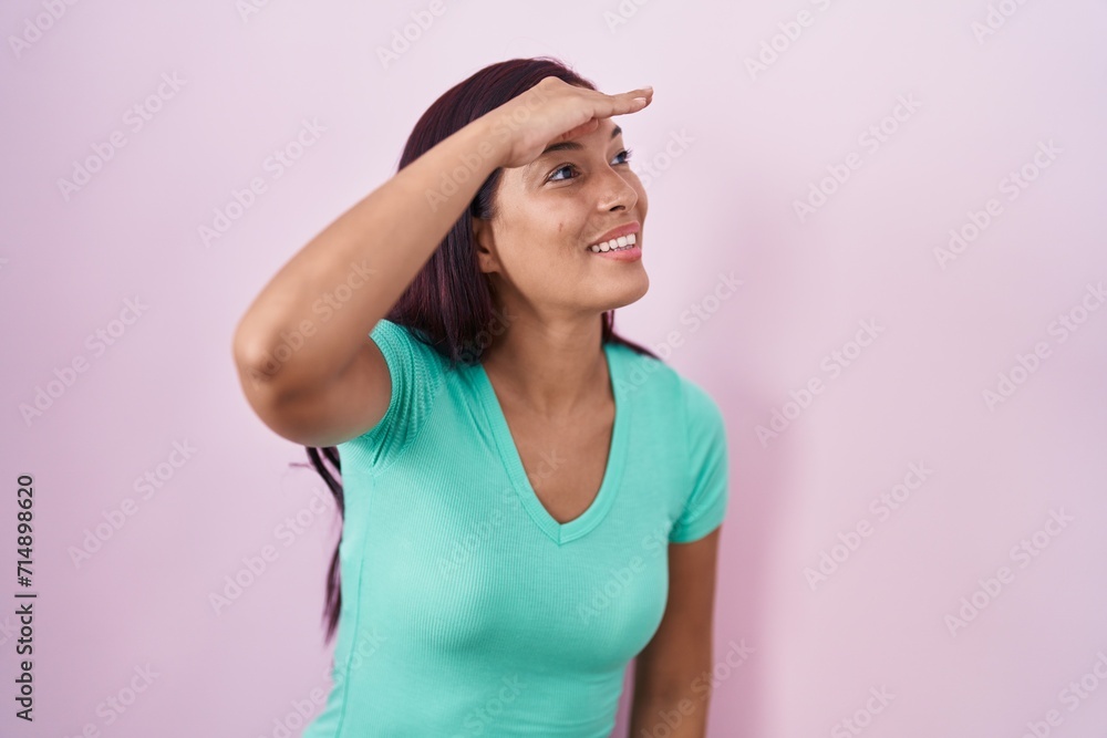 Young hispanic woman standing over pink background very happy and smiling looking far away with hand over head. searching concept. Young hispanic woman standing over pink background very happy and smiling looking far away with hand over head. searching concept.