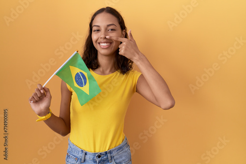 Young hispanic woman holding brazil flag pointing with hand finger to face and nose, smiling cheerful. beauty concept