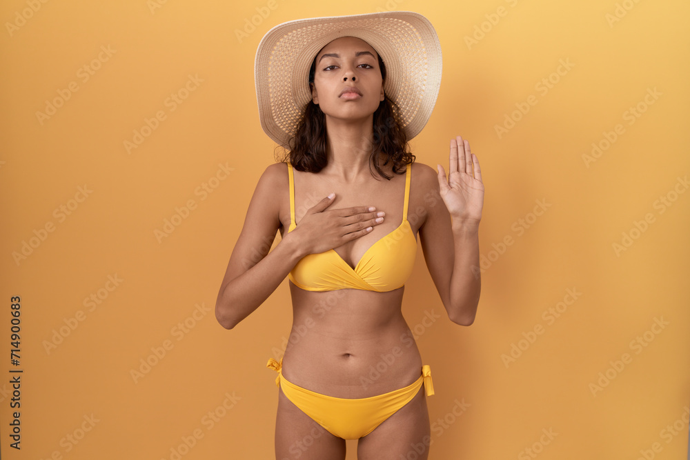 Young hispanic woman wearing bikini and summer hat swearing with hand on chest and open palm, making a loyalty promise oath Young hispanic woman wearing bikini and summer hat swearing with hand on chest and open palm, making a loyalty promise oath