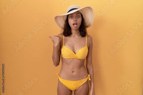 Young hispanic woman wearing bikini and summer hat surprised pointing with hand finger to the side  open mouth amazed expression.