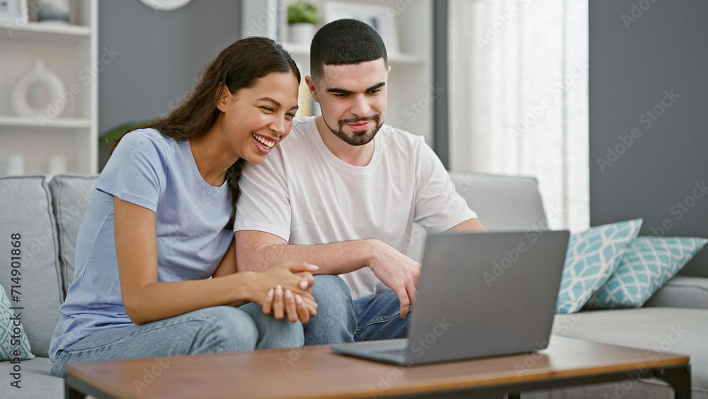 Cheerful, beautiful couple in love sitting together on a cozy sofa at home, smiling while using their laptop in their comfortable living room setting Cheerful, beautiful couple in love sitting together on a cozy sofa at home, smiling while using their laptop in their comfortable living room setting