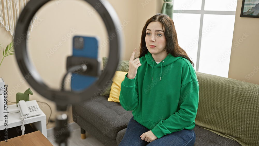 A young caucasian woman in a green hoodie gesturing indoors while recording with a smartphone and ring light in a cozy living room. A young caucasian woman in a green hoodie gesturing indoors while recording with a smartphone and ring light in a cozy living room.